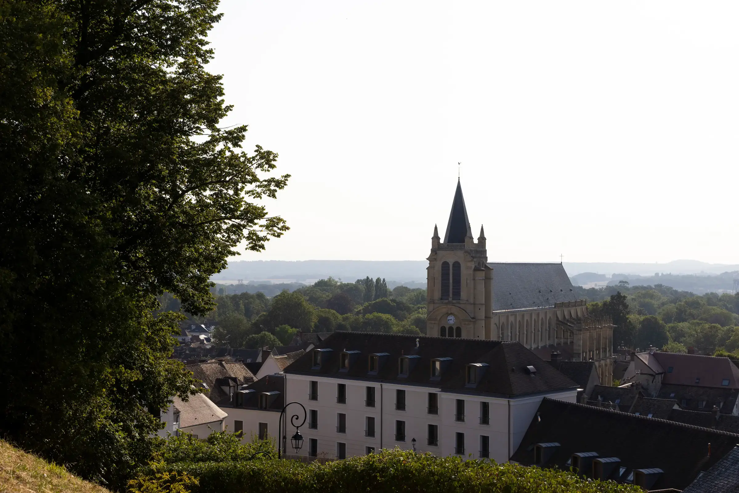 Une église gothique avec une flèche centrale et des toits d'ardoise, entourée de bâtiments résidentiels, vue à travers des arbres