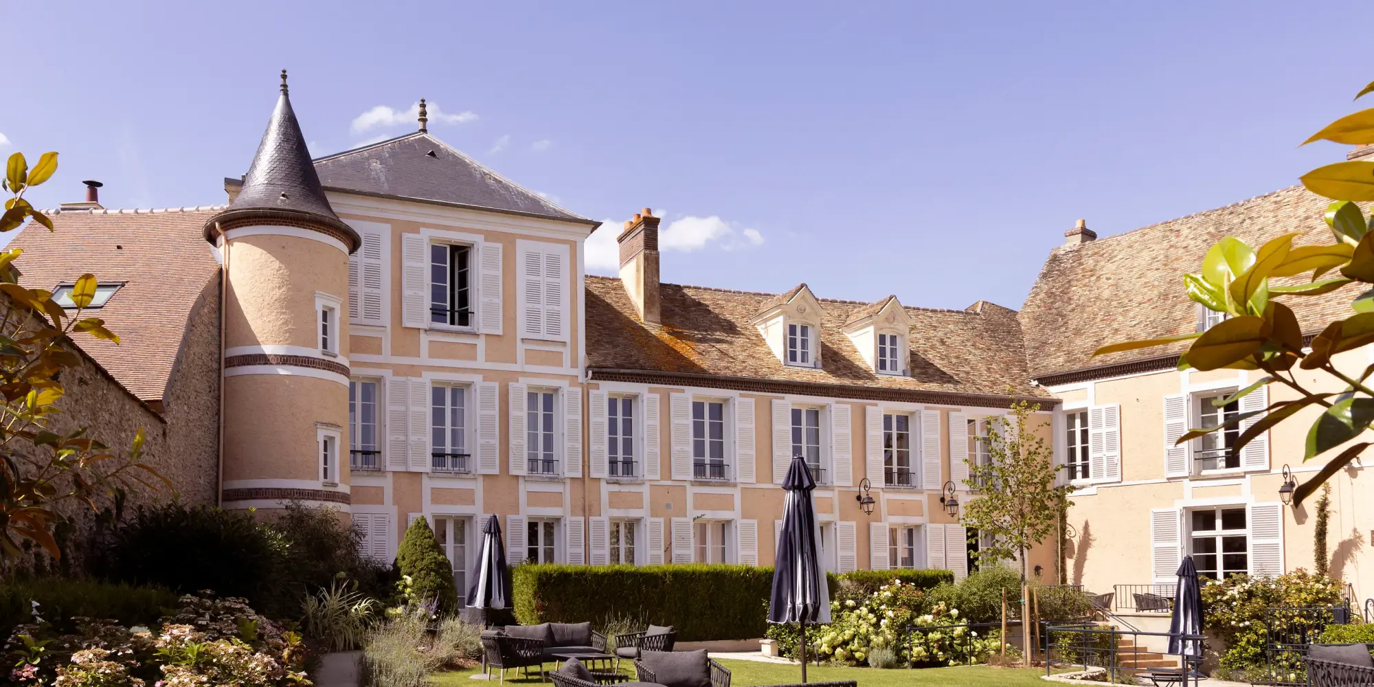 Un bâtiment historique avec une tour ronde et des fenêtres à volets blancs, entouré d'un jardin avec des chaises et des plantes