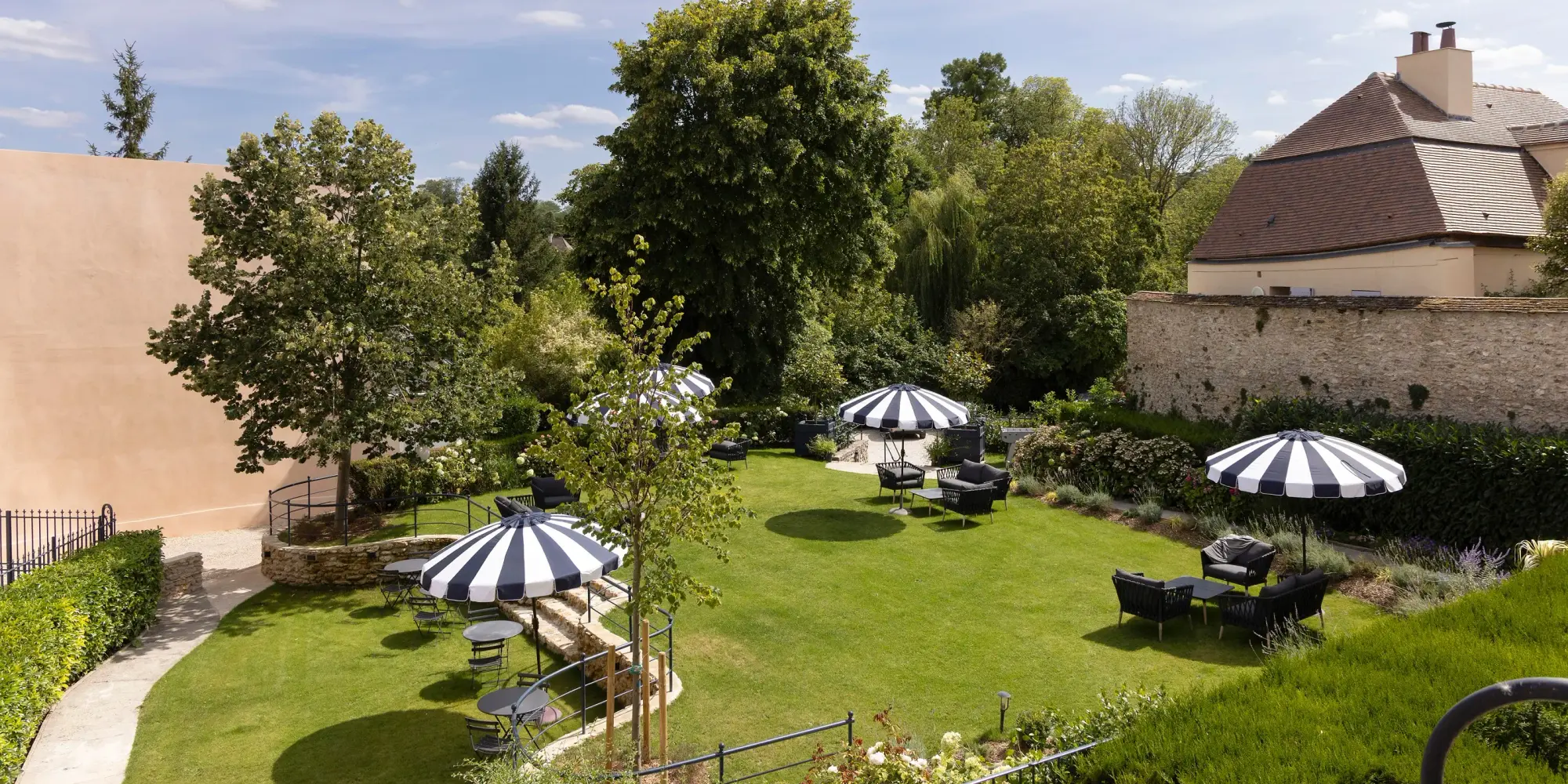 Jardin avec pelouse, arbres, plantes, meubles de jardin et parasols, entouré de bâtiments et d'une vieille muraille