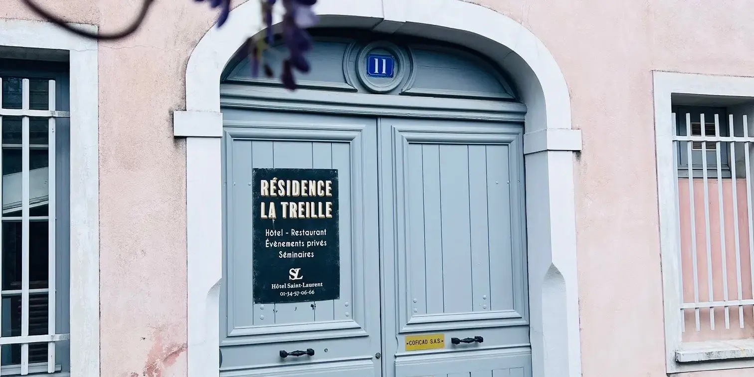 Porte d'entrée bleue d'un bâtiment rose avec une enseigne indiquant "Résidence La Treille" et des fleurs violettes en premier plan