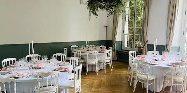 Salle de réception avec plusieurs tables rondes recouvertes de nappes blanches, des chaises assorties et des décorations florales suspendues au plafond