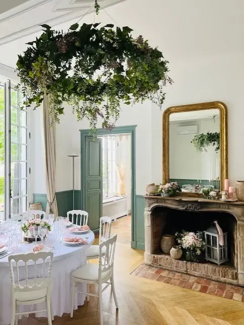 Salle à manger avec table dressée, cheminée décorée de fleurs et miroir, et une suspension florale au plafond