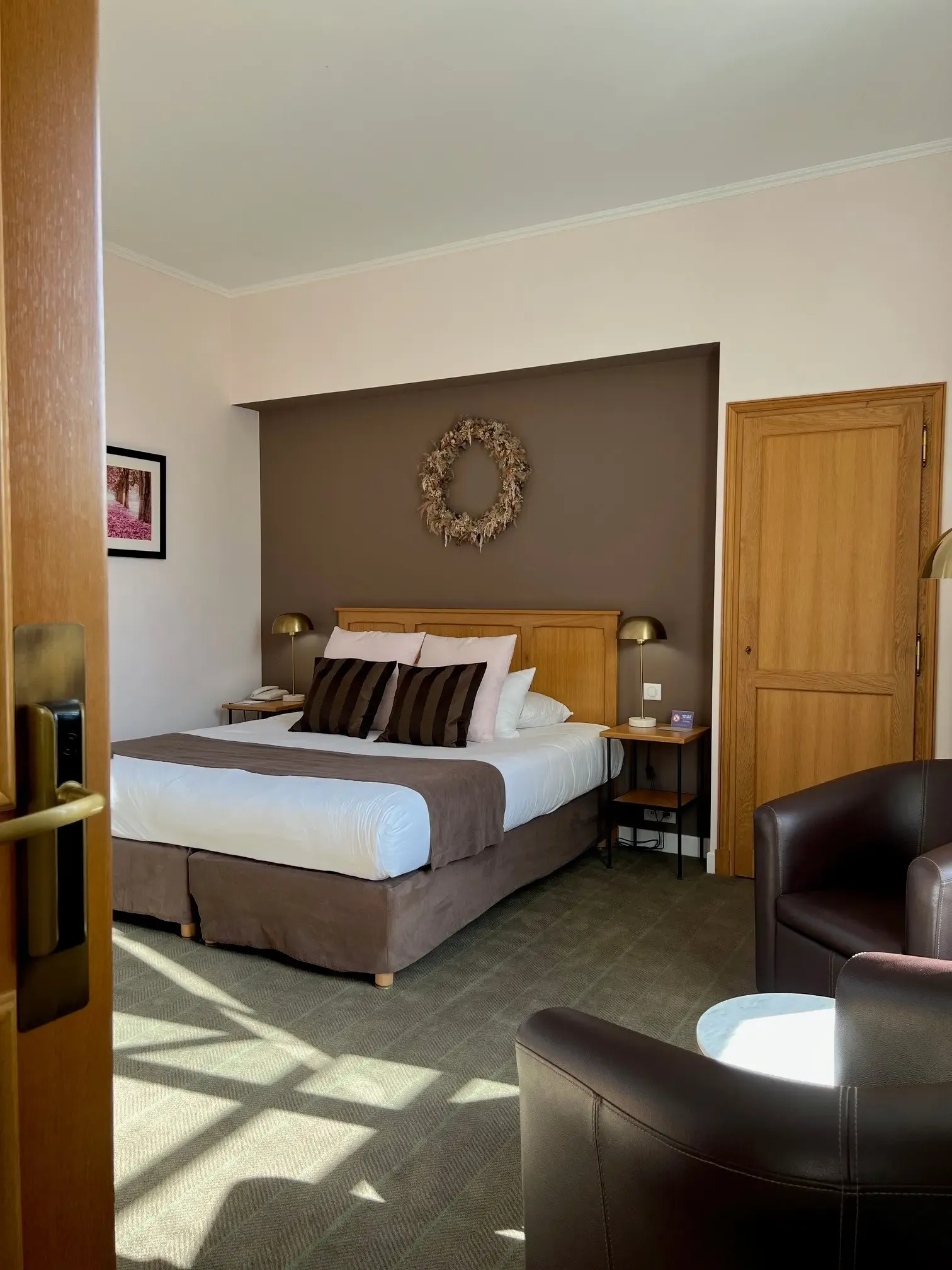 Chambre d'hôtel avec un lit double, une tête de lit en bois, deux tables de chevet avec lampes, un fauteuil en cuir et une porte en bois