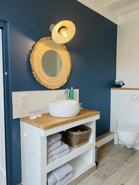 Un lavabo blanc sur un meuble en bois avec des serviettes, un panier et des produits de toilette, devant un miroir rond entouré d'un cadre en fibres naturelles, dans une salle de bain aux murs bleu foncé et au sol en carrelage clair