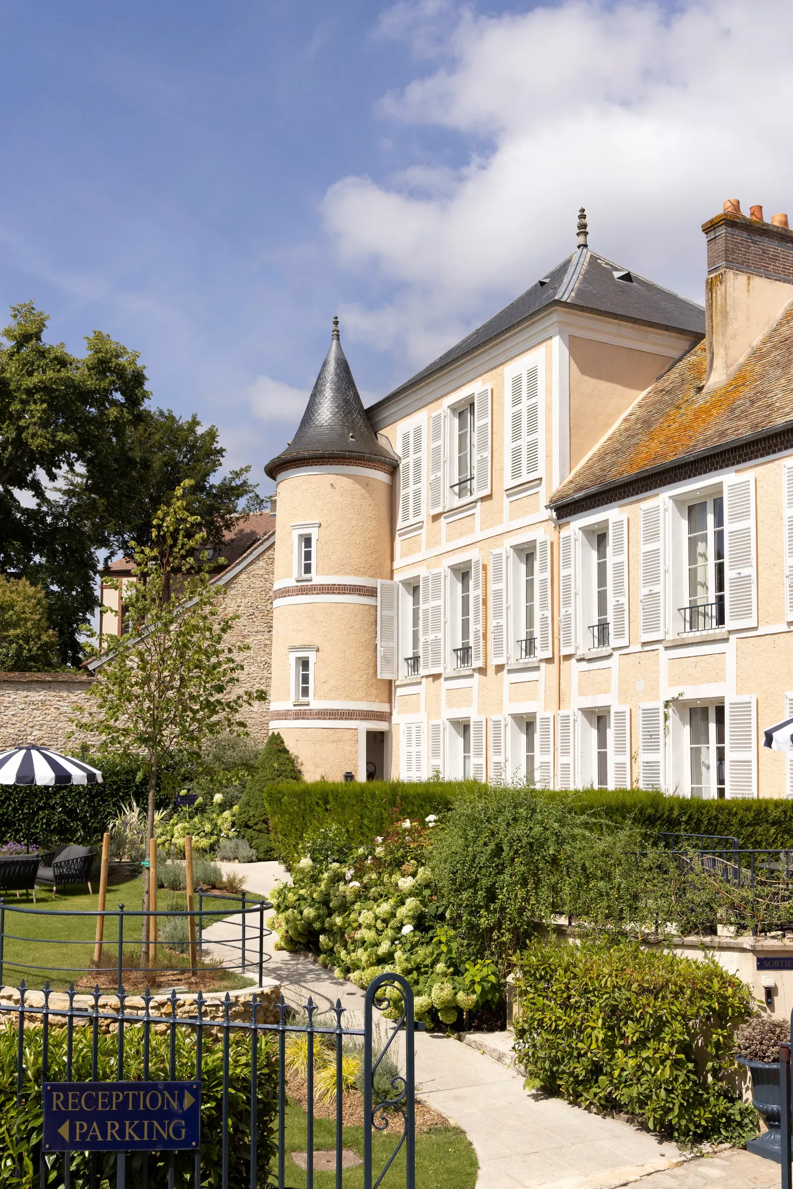 Un bâtiment ancien avec une tour ronde et des volets blancs, entouré d'un jardin avec des buissons et une allée menant à une grille marquée "Réception Parking"