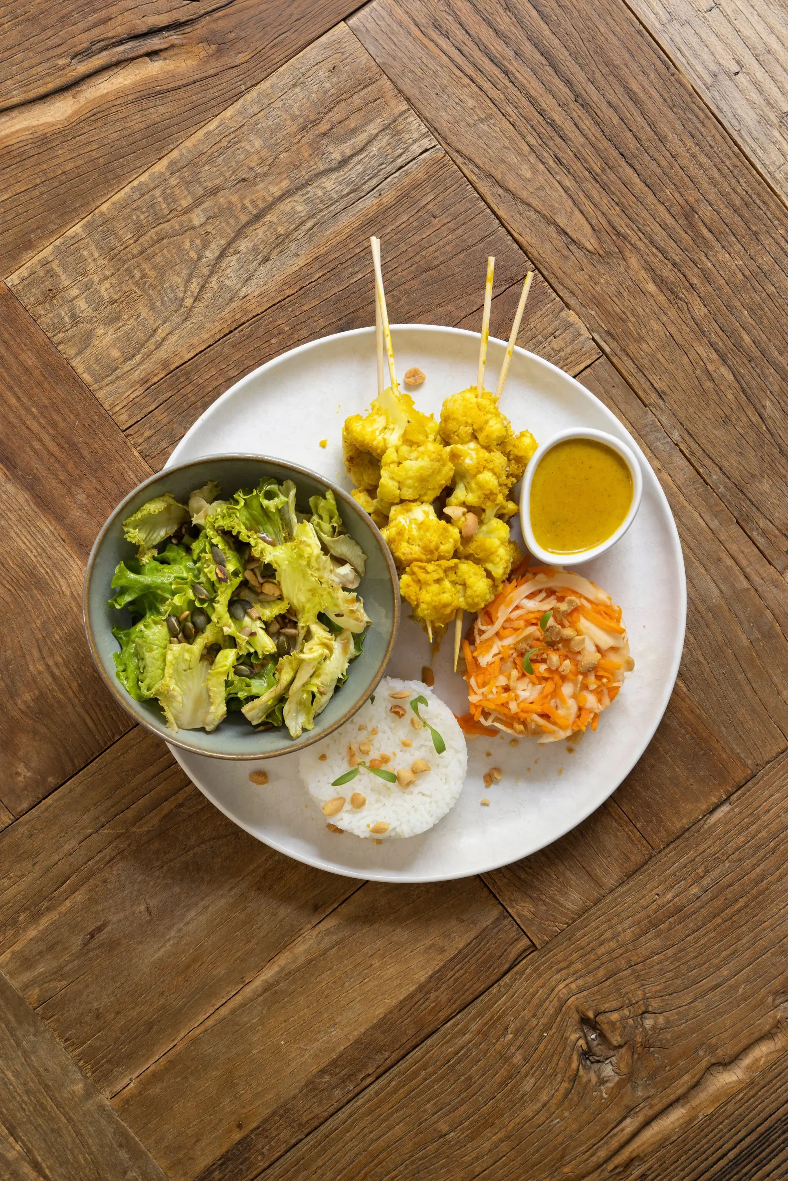 Un plat composé de brochettes de légumes frits, de riz, de salade et de carottes râpées, accompagné d'une sauce, présenté sur une table en bois