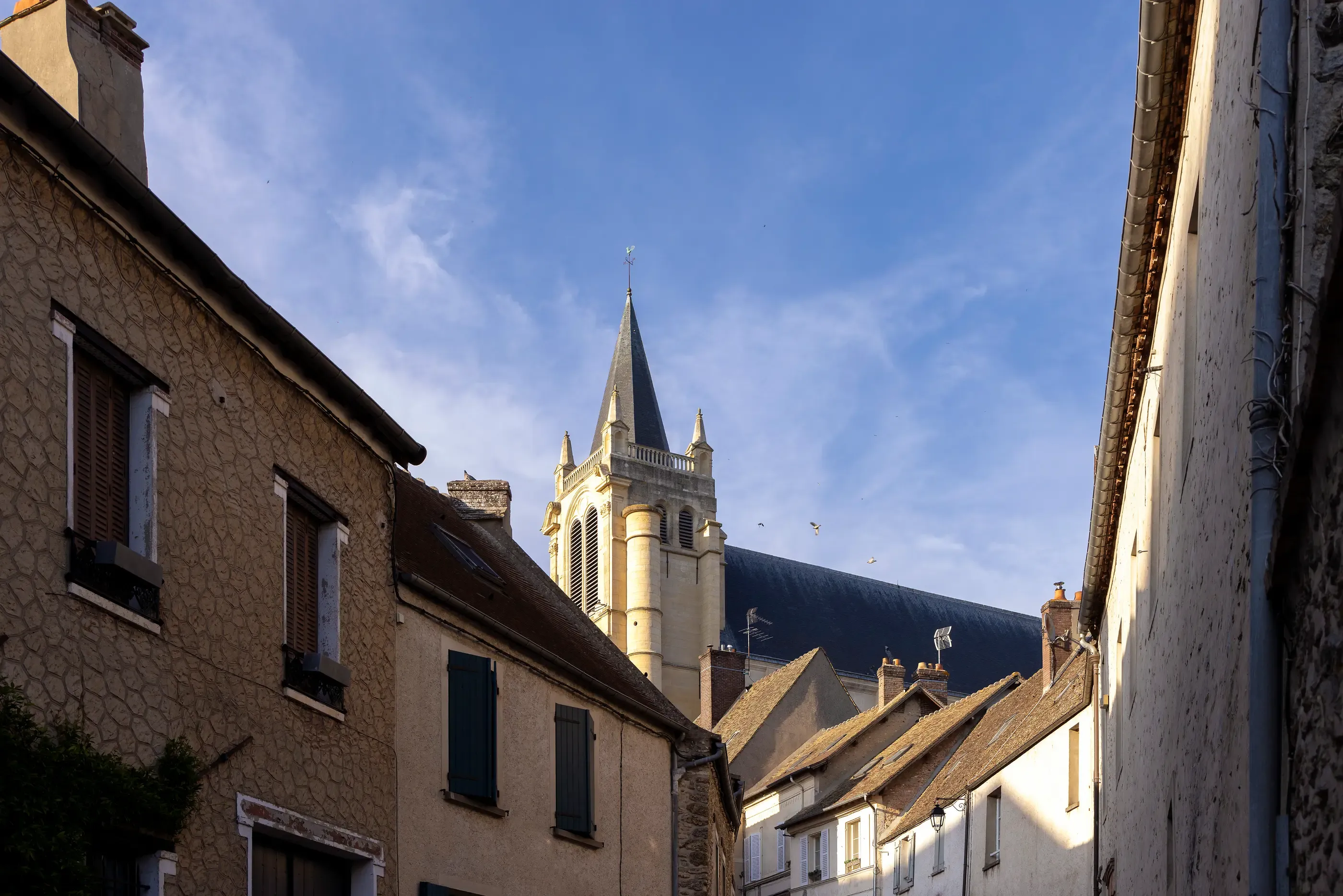Une rue étroite bordée de maisons anciennes avec un clocher gothique en arrière-plan sous un ciel partiellement nuageux