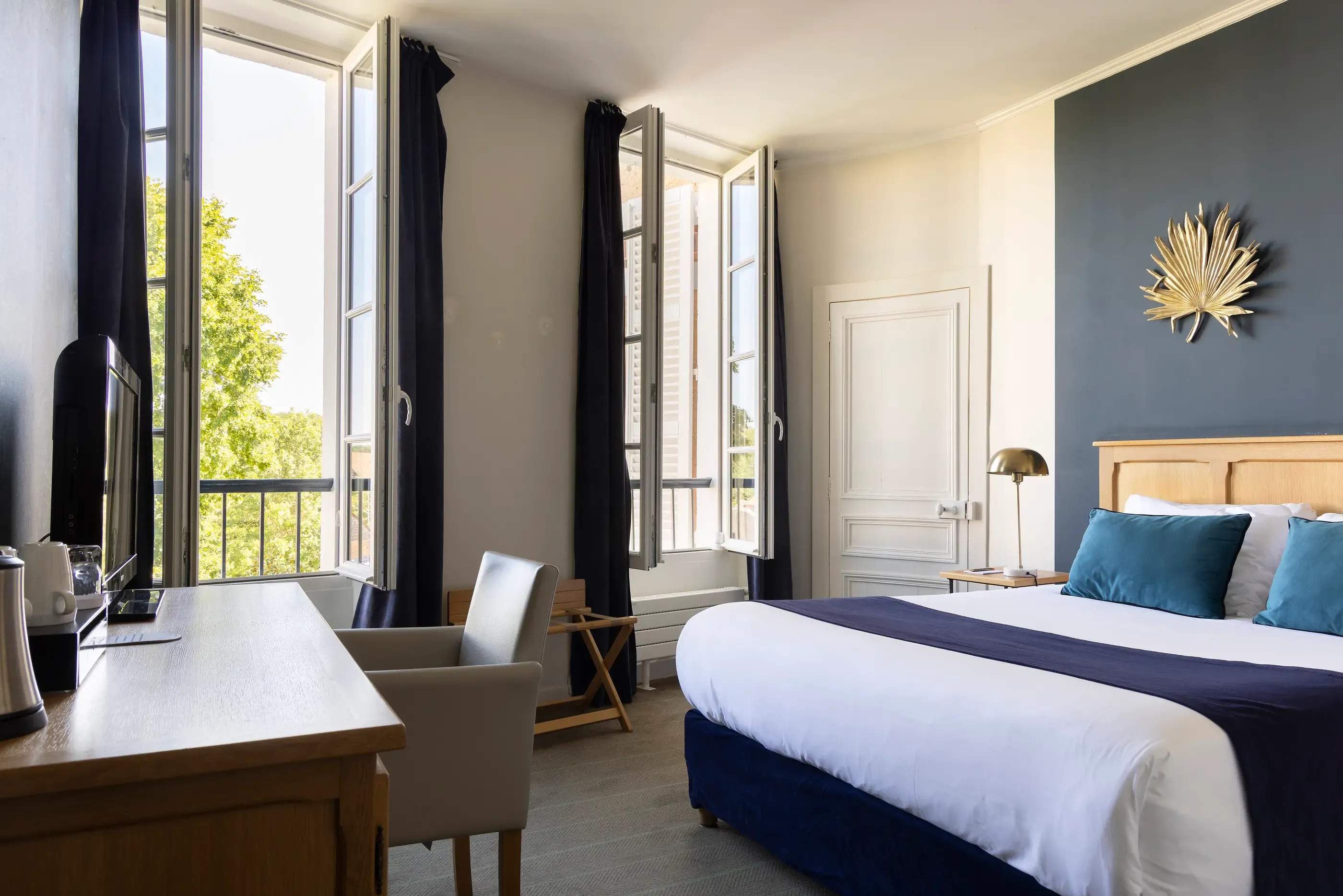 Chambre lumineuse avec lit double, table de travail, fauteuil et fenêtres ouvertes sur un balcon avec vue sur des arbres
