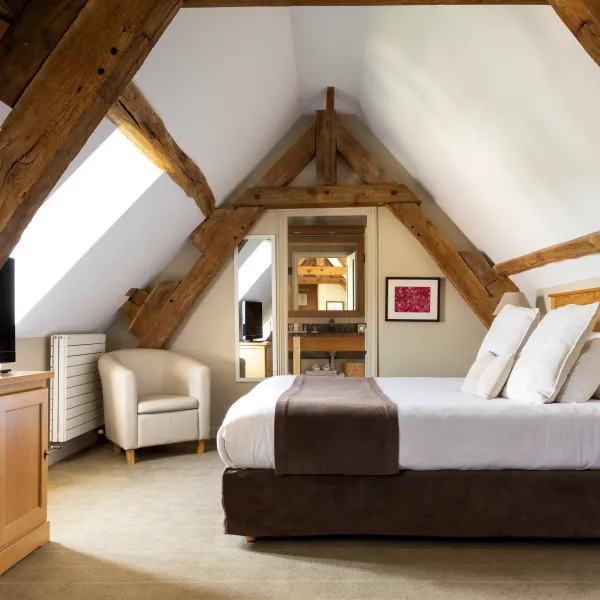 Chambre mansardée avec lit central, télévision, fauteuil et cheminée dans un cadre en bois apparent