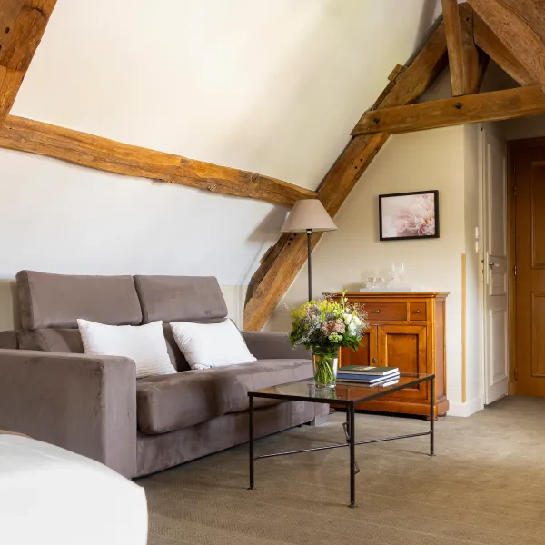 Un salon avec un canapé gris, une table basse, un vase de fleurs et des poufs, sous un plafond à poutres apparentes