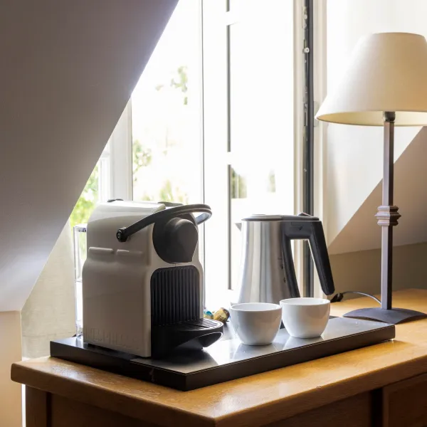 Un coin cuisine avec une machine à café, une bouilloire, deux tasses et une lampe sur un plan de travail en bois près d'une fenêtre