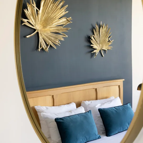 Reflet dans un miroir ovale doré d'un lit avec une tête de lit en bois clair, des coussins blancs et bleus, et deux décorations murales en forme de feuilles dorées sur un fond gris foncé