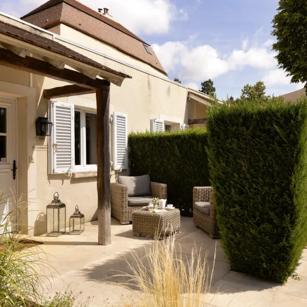Patio extérieur avec mobilier en osier, lanternes et haies taillées, adjacent à une maison aux volets fermés