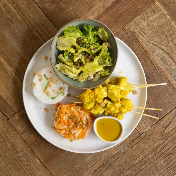 Un bol de salade verte, un tas de riz, des brochettes de légumes frits et une petite portion de carottes râpées sur une assiette blanche posée sur une table en bois
