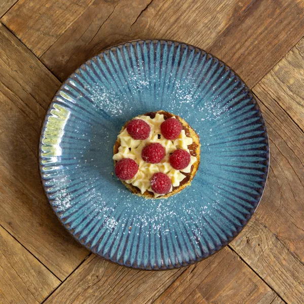 Petite tarte garnie de crème et de framboises sur une assiette bleue à bord festonné posée sur une table en bois