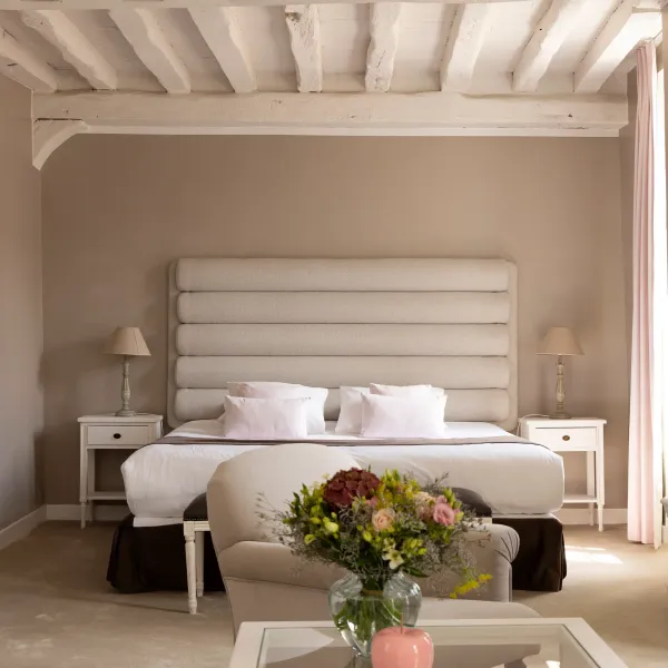 Chambre avec lit double, tête de lit en tissu, deux tables de chevet avec lampes, un fauteuil, une table basse avec un vase de fleurs et une plante accrochée au mur